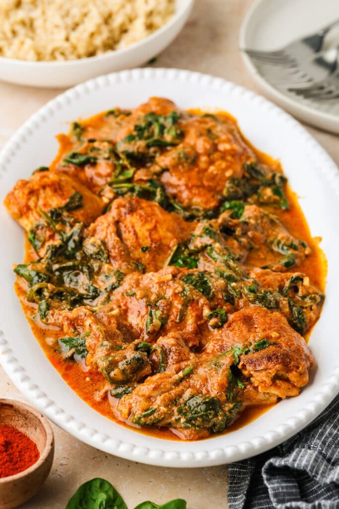 Oval white platter filled with braised chicken thighs in a rich, orange-red tomato sauce with wilted spinach and herbs. Plates, rice, and a bowl of spice are visible in the background.