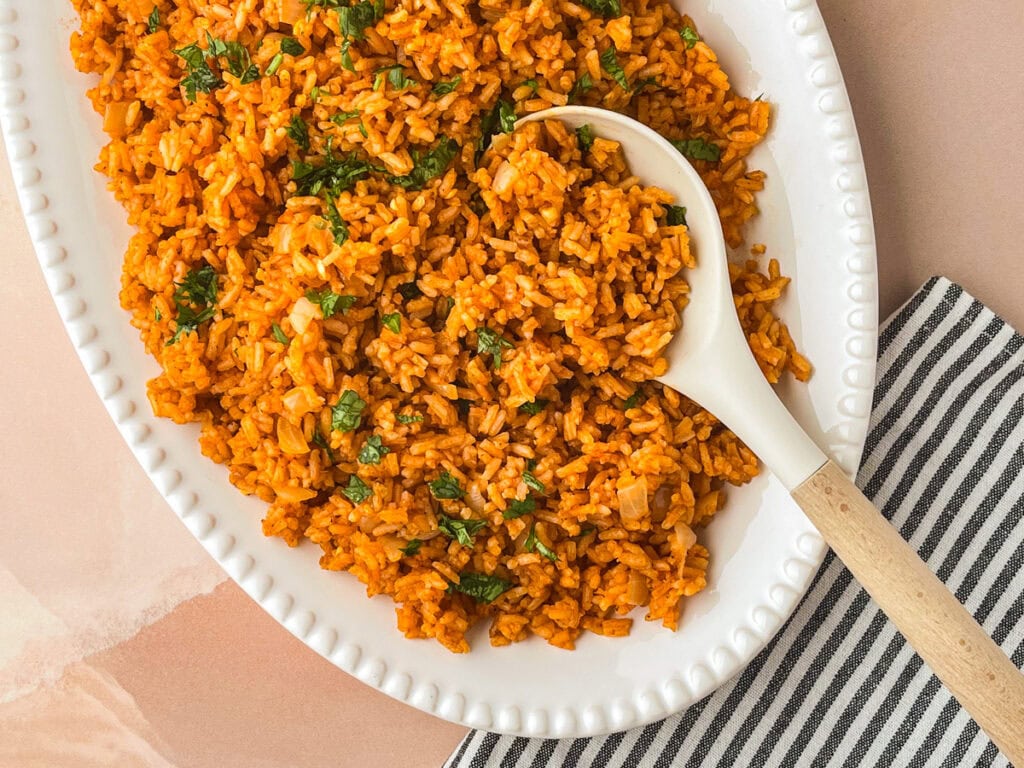 A white oval dish filled with Mexican rice, garnished with chopped herbs, and a white serving spoon resting in the rice. The dish sits on a striped black and white napkin.