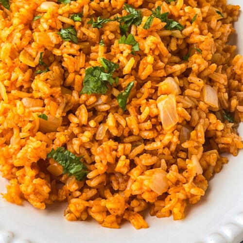 A close-up of a plate of Mexican rice, featuring orange-colored rice mixed with chopped onions and garnished with fresh cilantro. The dish is served on a white plate with a decorative edge.
