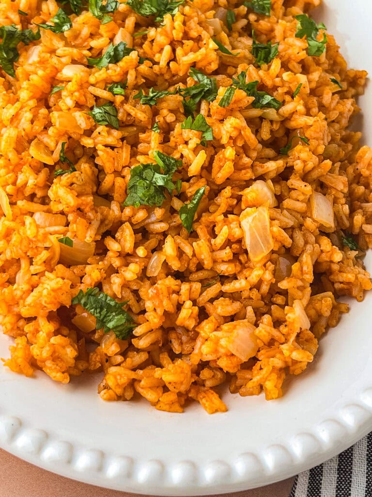 A close-up of a plate of Mexican rice, featuring orange-colored rice mixed with chopped onions and garnished with fresh cilantro. The dish is served on a white plate with a decorative edge.