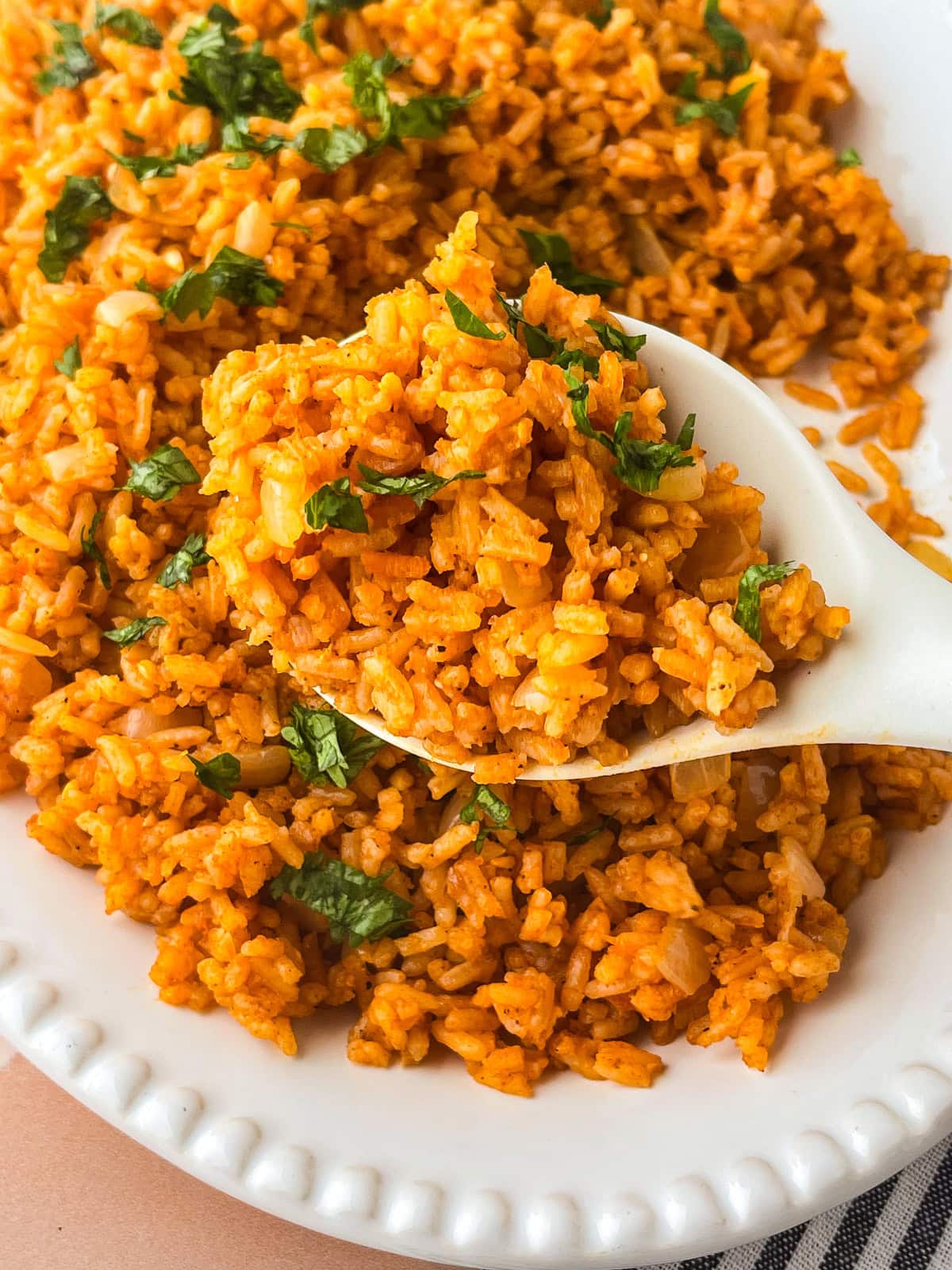 A close-up of a white spoon scooping seasoned orange rice garnished with chopped fresh cilantro from a white dish.