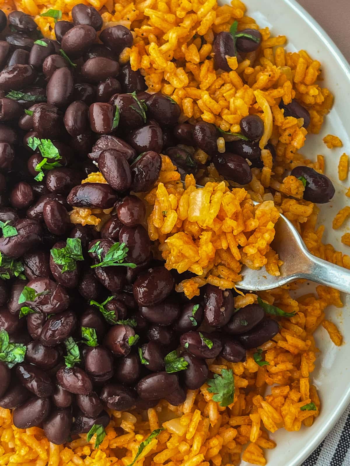 A close-up of a plate filled with orange Mexican rice and black beans, garnished with chopped cilantro. A fork is scooping up some of the seasoned rice from the plate.
