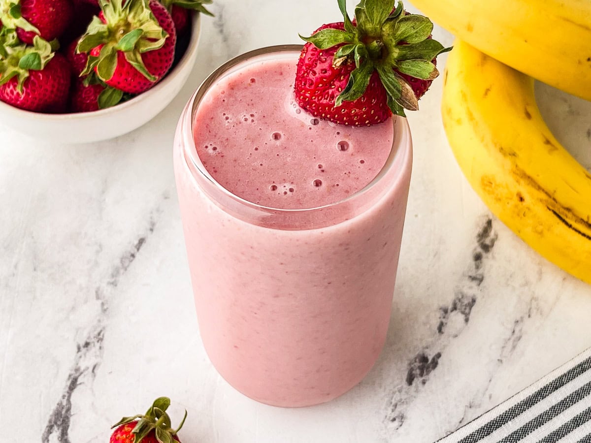 A glass of pink strawberry banana smoothie garnished with a whole strawberry, placed on a marble surface next to fresh strawberries in a bowl and ripe bananas.