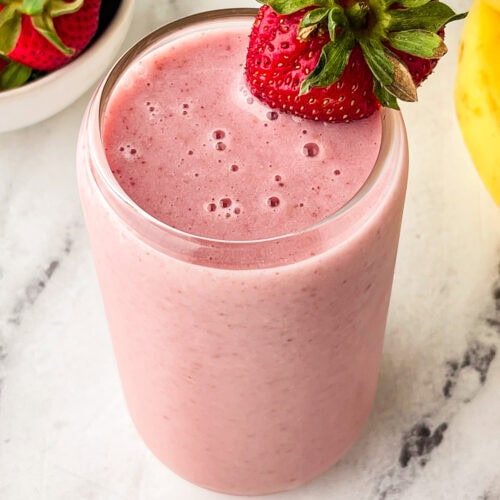 A glass filled with pink strawberry banana smoothie, garnished with a whole strawberry on the rim, sits on a marble surface next to fresh strawberries and a bunch of bananas.