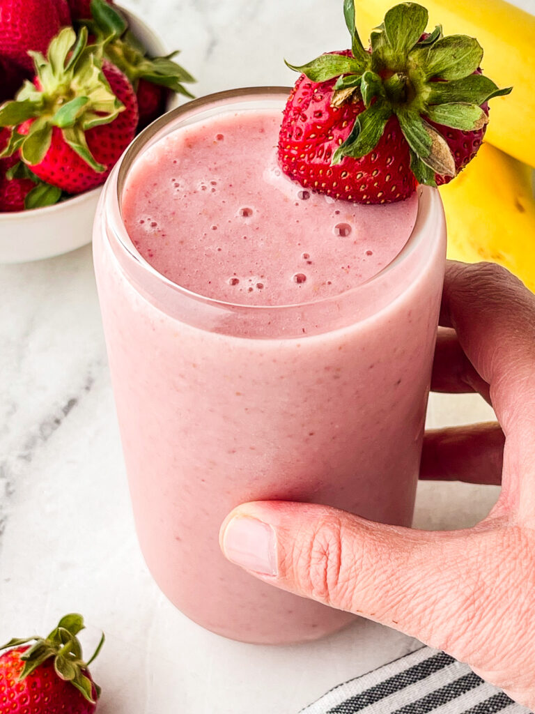 A hand holds a glass of pink strawberry smoothie garnished with a whole strawberry. Fresh strawberries and a banana are visible in the background on a white surface.