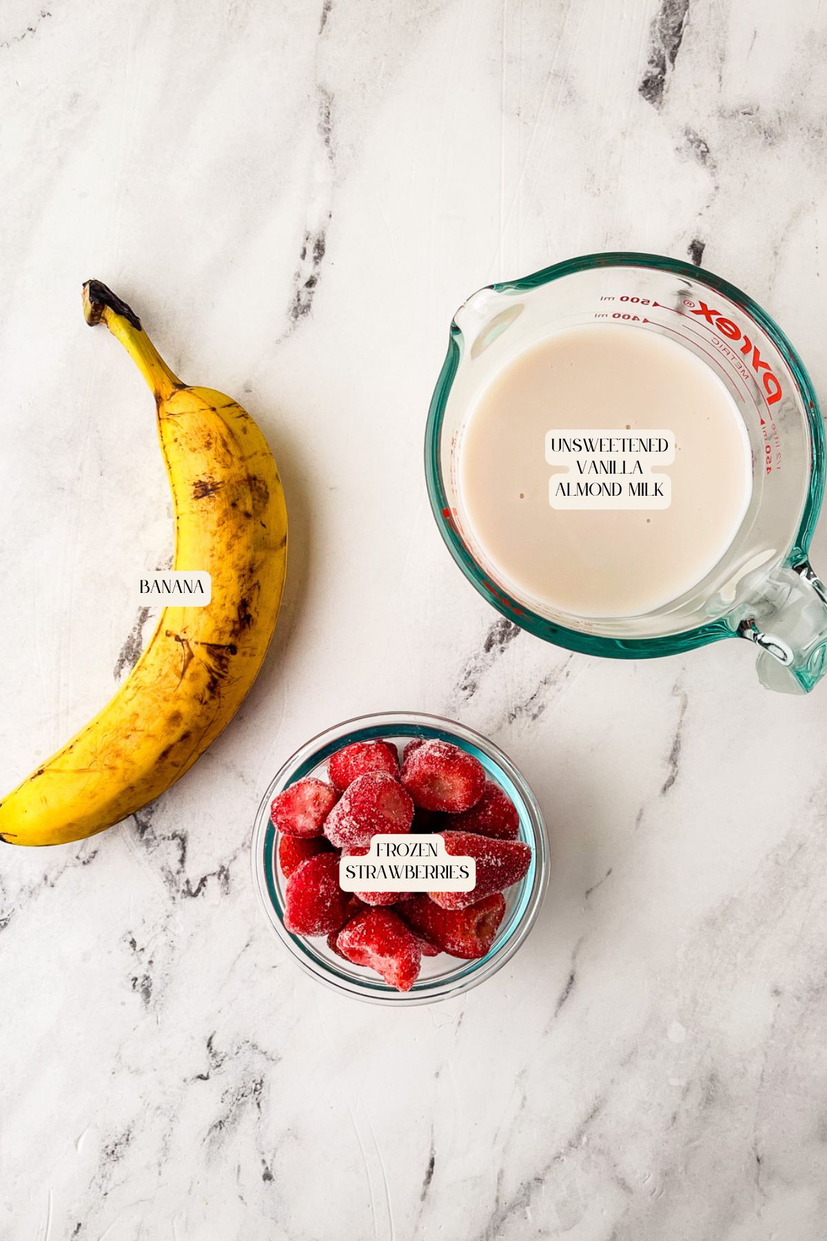 A banana, a measuring cup of unsweetened vanilla almond milk, and a bowl of frozen strawberries are arranged on a white marble surface. Each item is labeled.