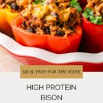 Colorful red and orange bison stuffed peppers are filled with a savory black bean mixture, topped with melted cheese and fresh herbs, and arranged in a baking dish. Text overlay reads: “High Protein Bison Stuffed Peppers.”.