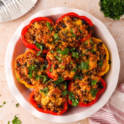 A white dish filled with baked stuffed bell peppers topped with melted cheese and chopped herbs, surrounded by a plate with forks, a small bowl of herbs, and a striped kitchen towel.