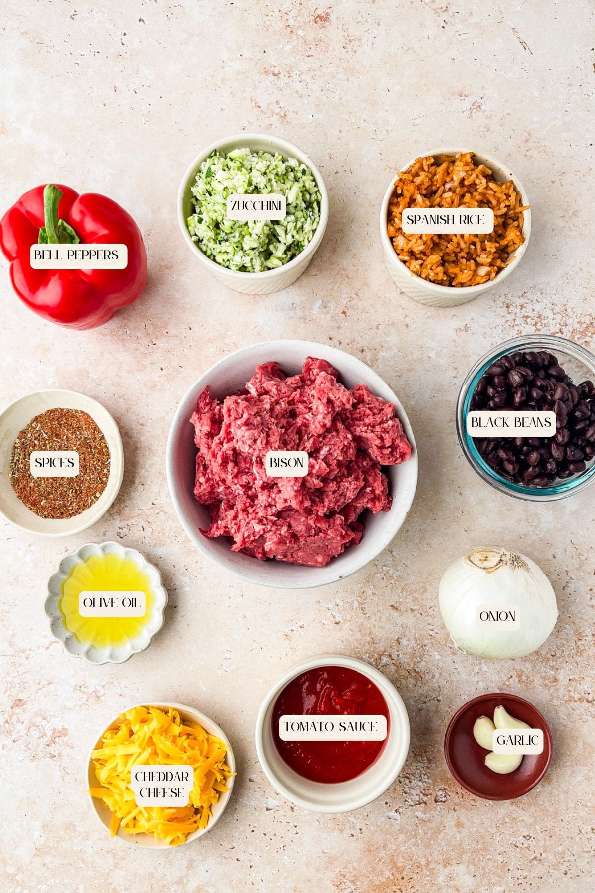 Top-down view of ingredients in bowls for a recipe: chopped bell peppers, zucchini, Spanish rice, black beans, spices, olive oil, grated cheddar cheese, tomato sauce, chopped onion, garlic, and ground bison.