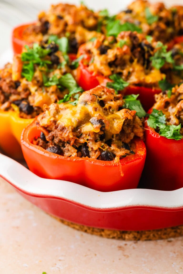 A close-up of stuffed bell peppers filled with a mixture of ground meat, black beans, rice, melted cheese, and topped with fresh chopped herbs, arranged in a red baking dish.
