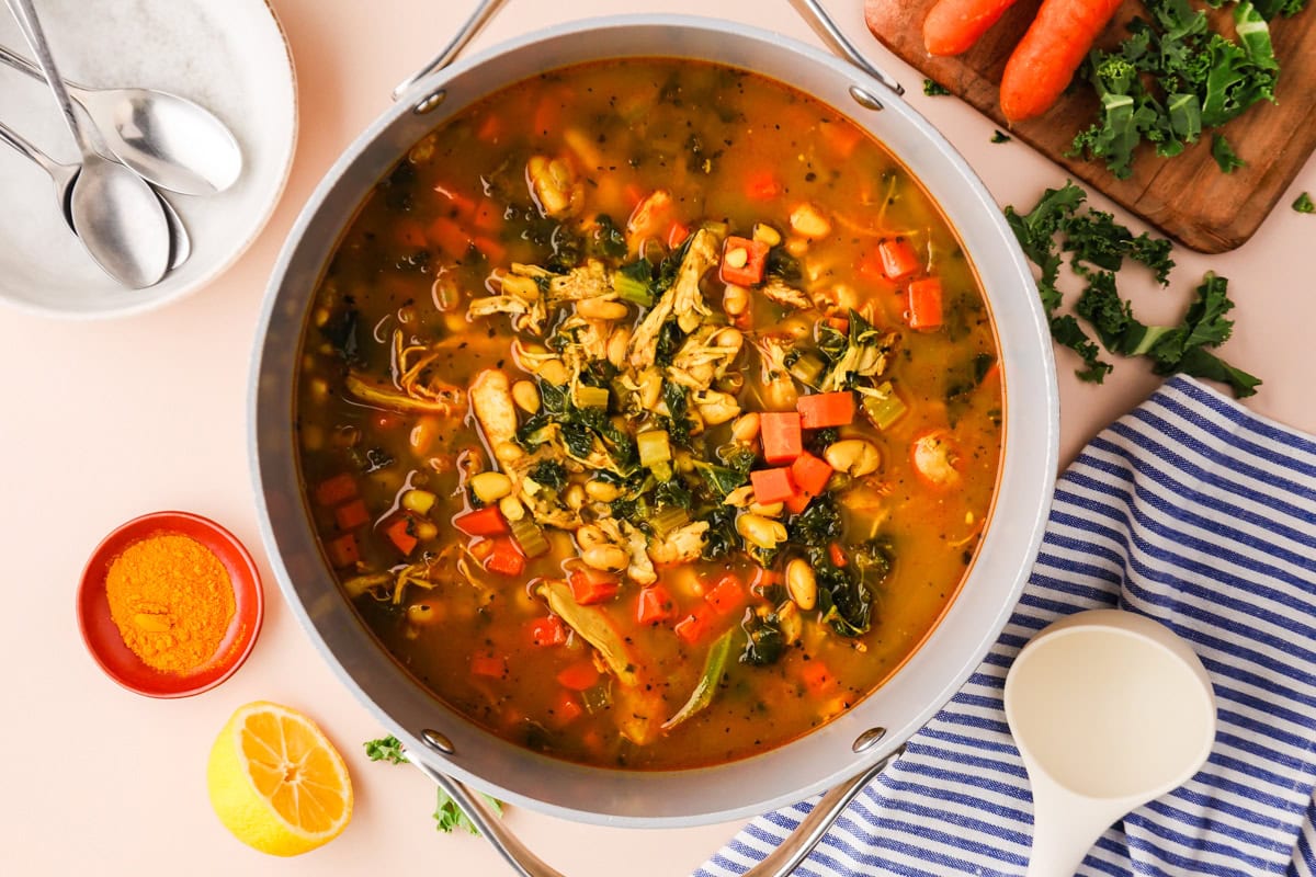 Large pot of colorful vegetable soup with shredded chicken, carrots, greens, and beans. Surrounding items include turmeric, lemon, striped towel, spoon, and cutting board with carrots and kale.