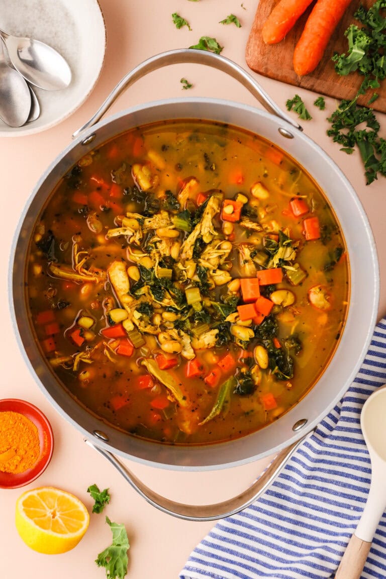 A pot of hearty vegetable and chicken soup with carrots, leafy greens, beans, and broth sits on a light surface, surrounded by sliced carrots, fresh herbs, a bowl of turmeric, a lemon half, and a striped kitchen towel.
