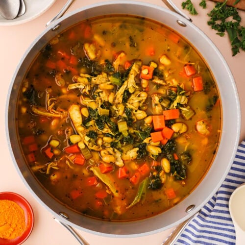 A pot of hearty vegetable soup with shredded chicken, carrots, beans, greens, and herbs sits on a beige surface, surrounded by spoons, raw carrots, chopped herbs, a lemon half, and a small bowl of turmeric powder.