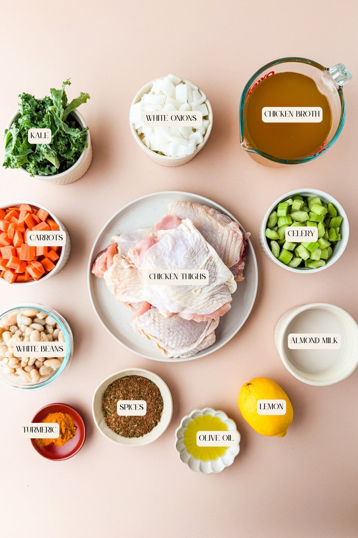 A flat lay of chicken and white bean soup ingredients: raw chicken thighs in the center, surrounded by bowls of kale, carrots, celery, white beans, almond milk, onions, spices, turmeric, olive oil, a lemon, and chicken broth.