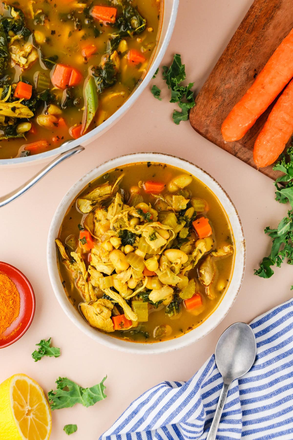 A bowl of vegetable soup with chickpeas, carrots, leafy greens, and yellow broth sits next to a pot of soup, fresh carrots, a lemon half, a striped towel, and a spoon on a light surface.