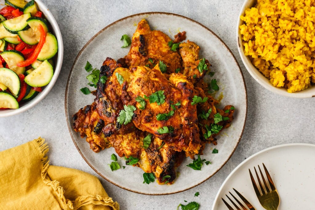 A plate of grilled, Greek yogurt chicken garnished with fresh herbs is surrounded by a bowl of yellow rice, a bowl of mixed vegetable salad, and a plate with two forks and a yellow napkin on a light surface.