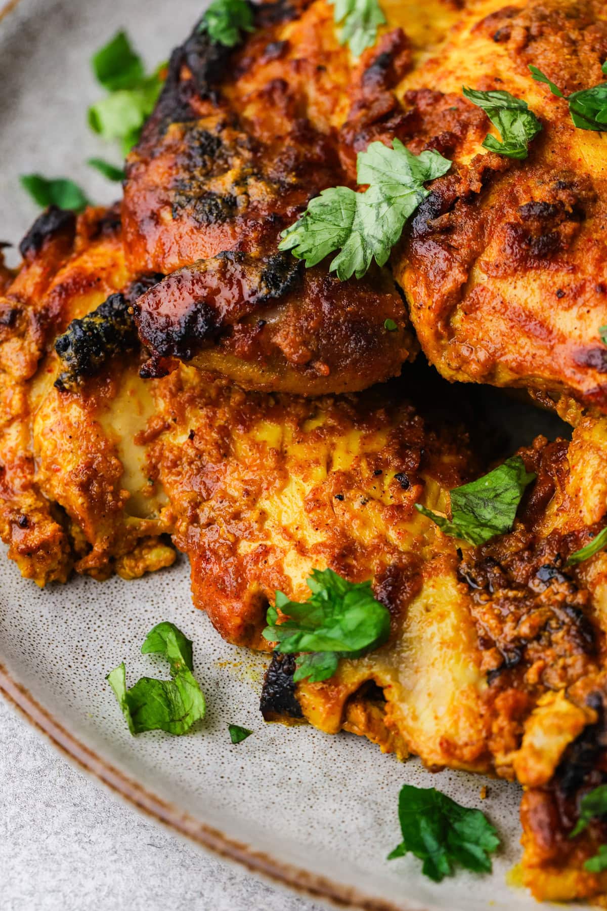 Close-up of flavorful, marinated greek yogurt chicken pieces garnished with fresh cilantro on a ceramic plate. The chicken is charred in spots and coated with a rich, orange-red spice mixture.
