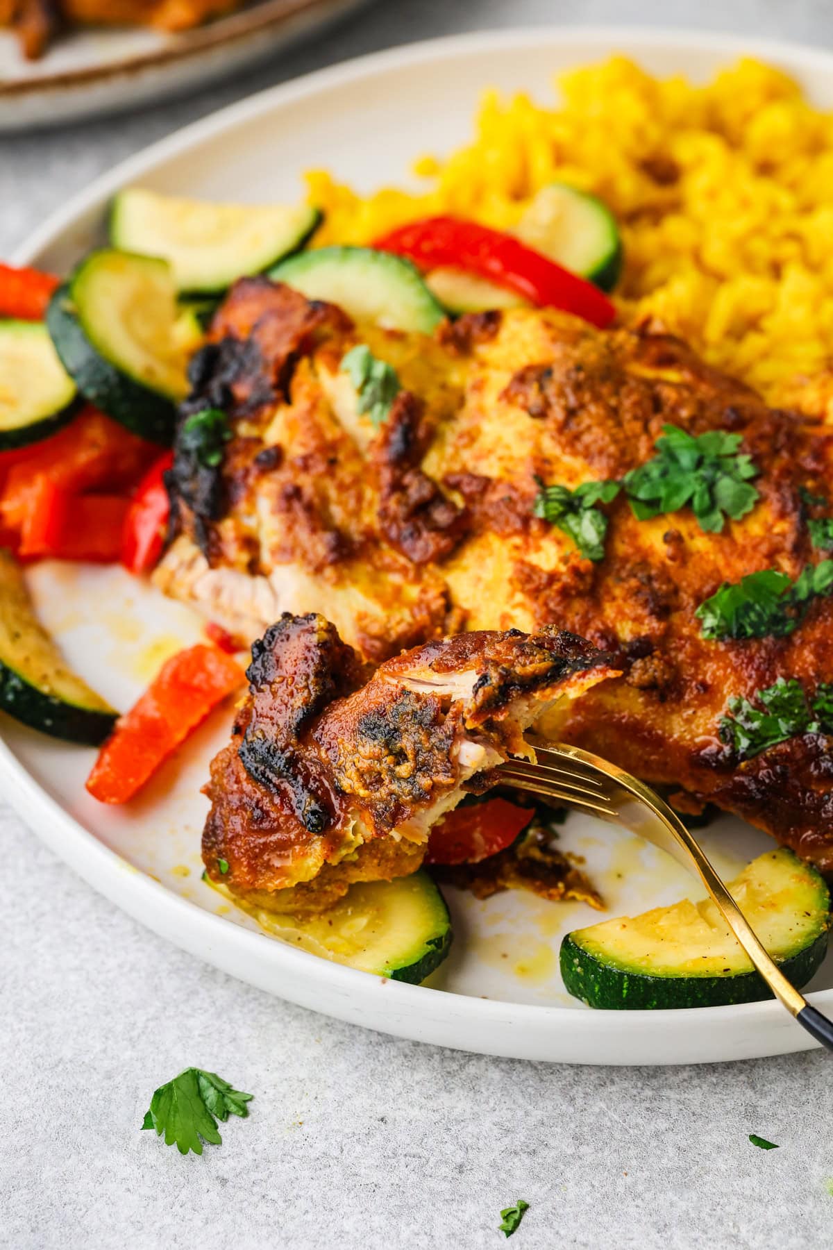 A close-up of a plate with grilled, seasoned Greek yogurt chicken, yellow rice, and sautéed vegetables including zucchini and red bell pepper. A fork holds a bite of chicken, and fresh herbs are sprinkled on top.