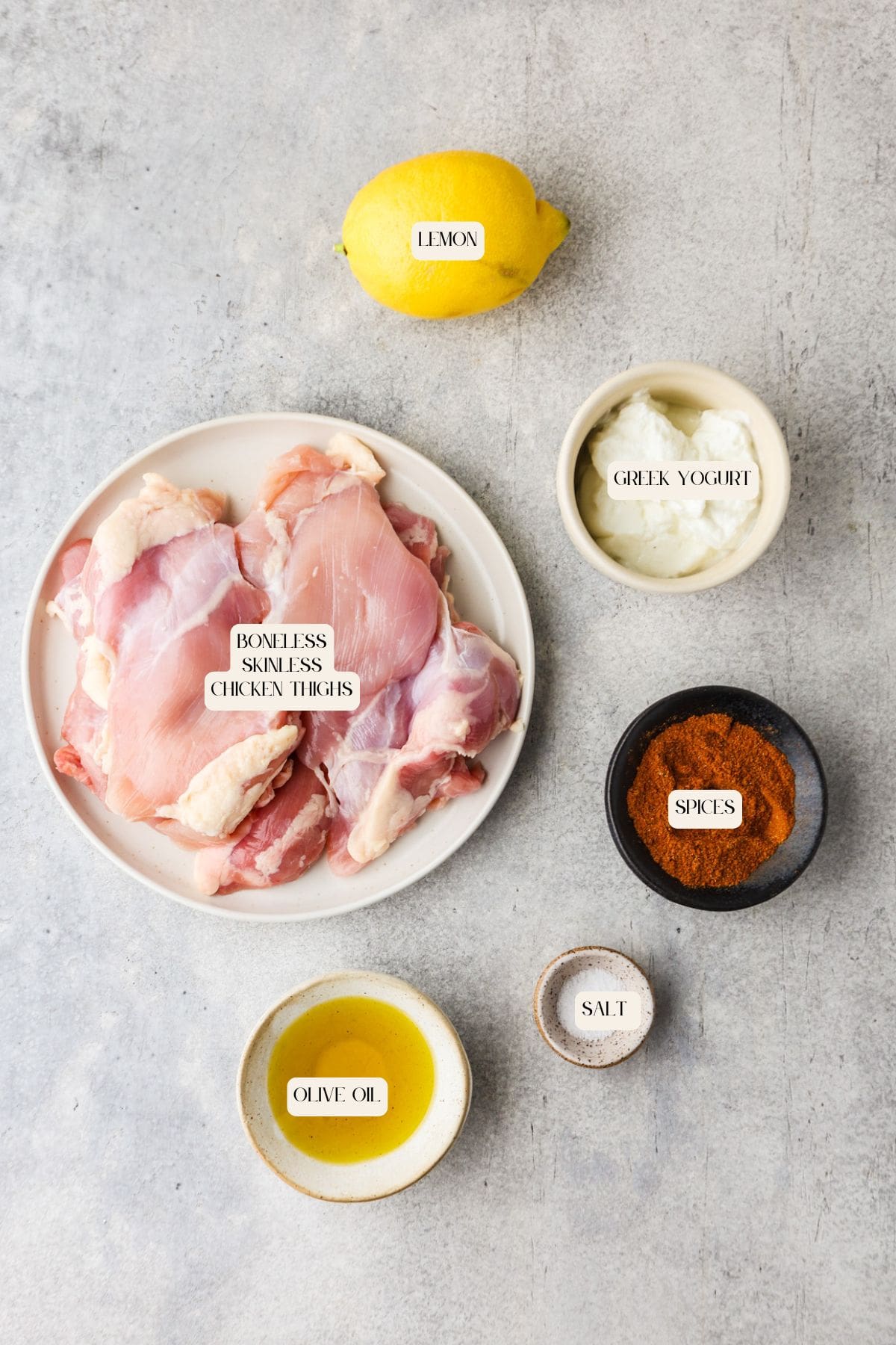 A flat lay of ingredients on a gray surface for greek yogurt chicken: a plate of boneless skinless chicken thighs, a lemon, Greek yogurt, spices, olive oil, and salt. Each is labeled for easy prep.