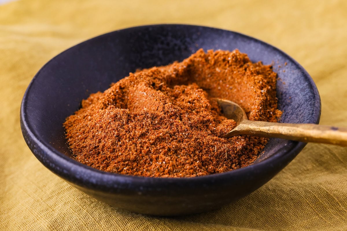 A black bowl filled with a pile of reddish-brown spice mix, with a small golden spoon resting in the spices, set on a yellow cloth background.