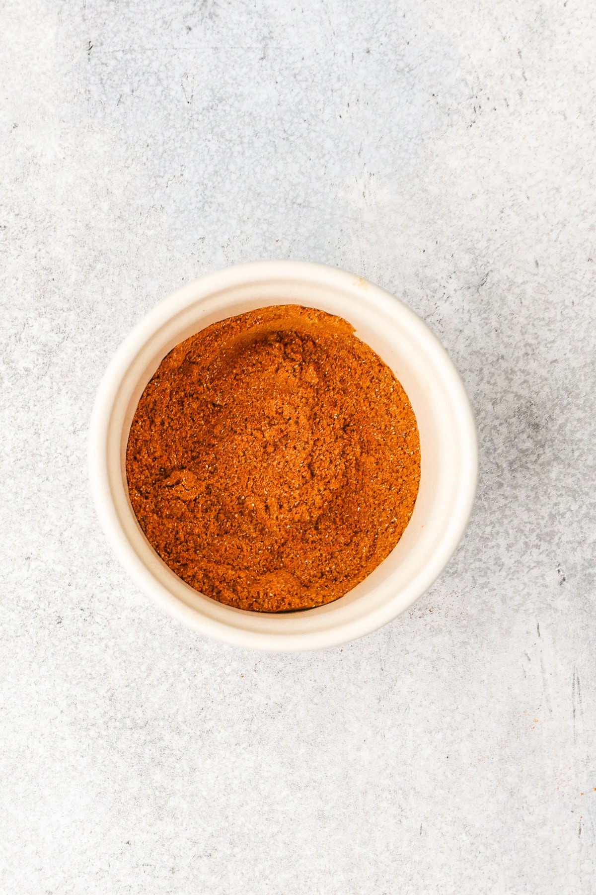 A small white bowl filled with a finely ground reddish-brown spice mixture, placed on a light gray textured surface.