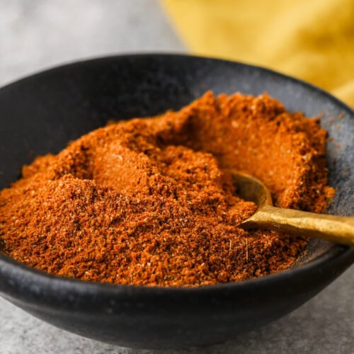 A black bowl filled with a reddish-brown spice blend, with a small gold spoon resting in the mixture, sits on a light gray surface. A yellow cloth is in the background.