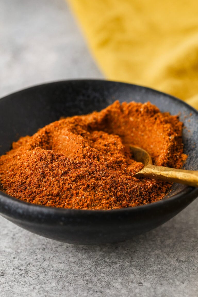 A black bowl filled with a reddish-brown spice blend, with a small gold spoon resting in the mixture, sits on a light gray surface. A yellow cloth is in the background.