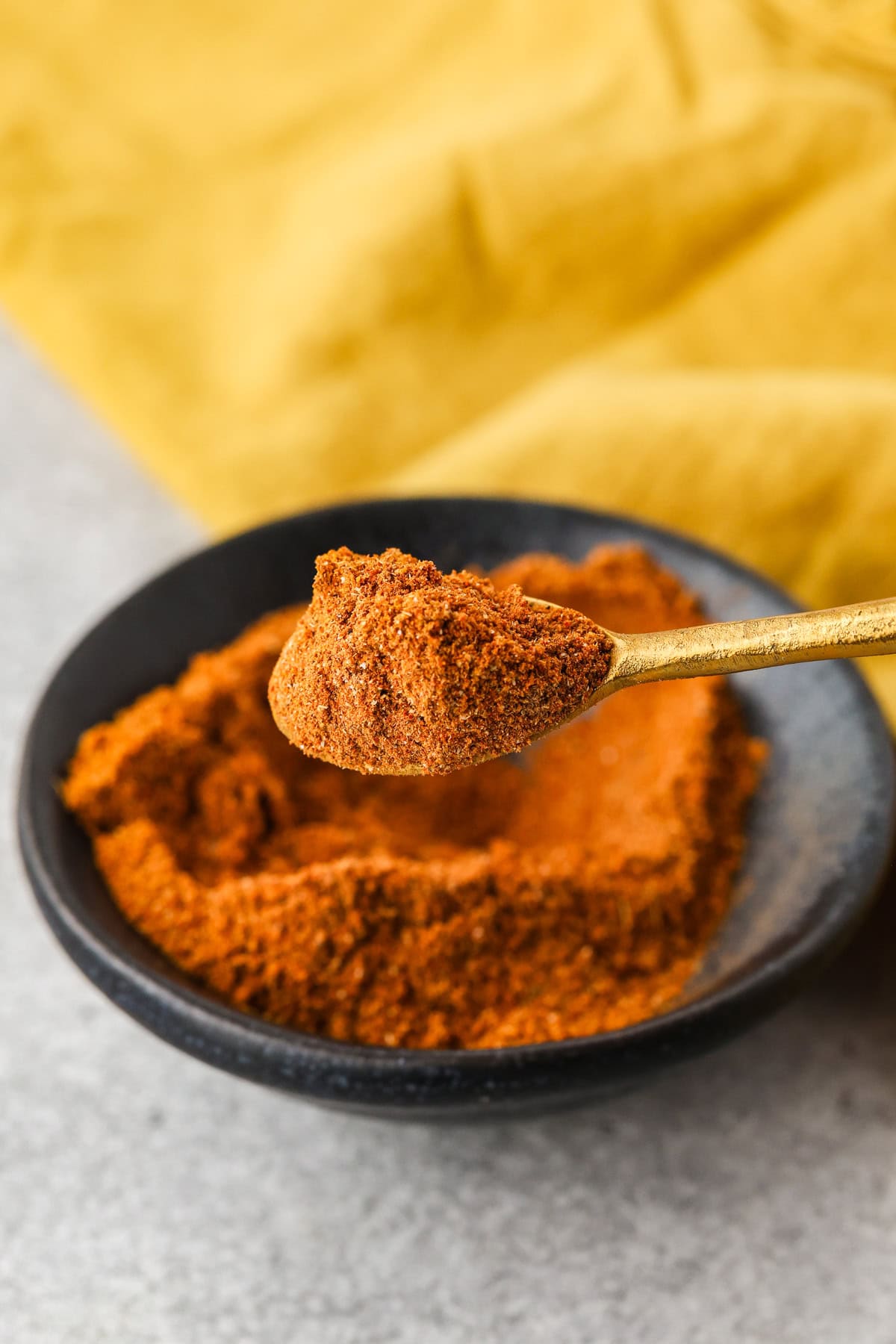 A gold spoon holding a heap of reddish-brown spice blend is held above a black bowl filled with more of the same spice, with a yellow cloth in the background.