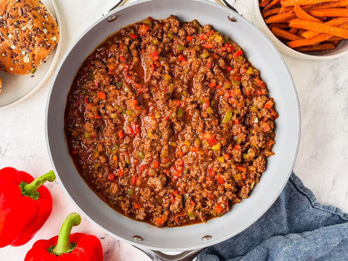 A skillet filled with cooked sloppy joe mixture made of ground beef, red and green bell peppers, and sauce sits on a white surface. Nearby are two fresh red bell peppers, a bowl of sweet potato fries, burger buns, and a blue napkin.