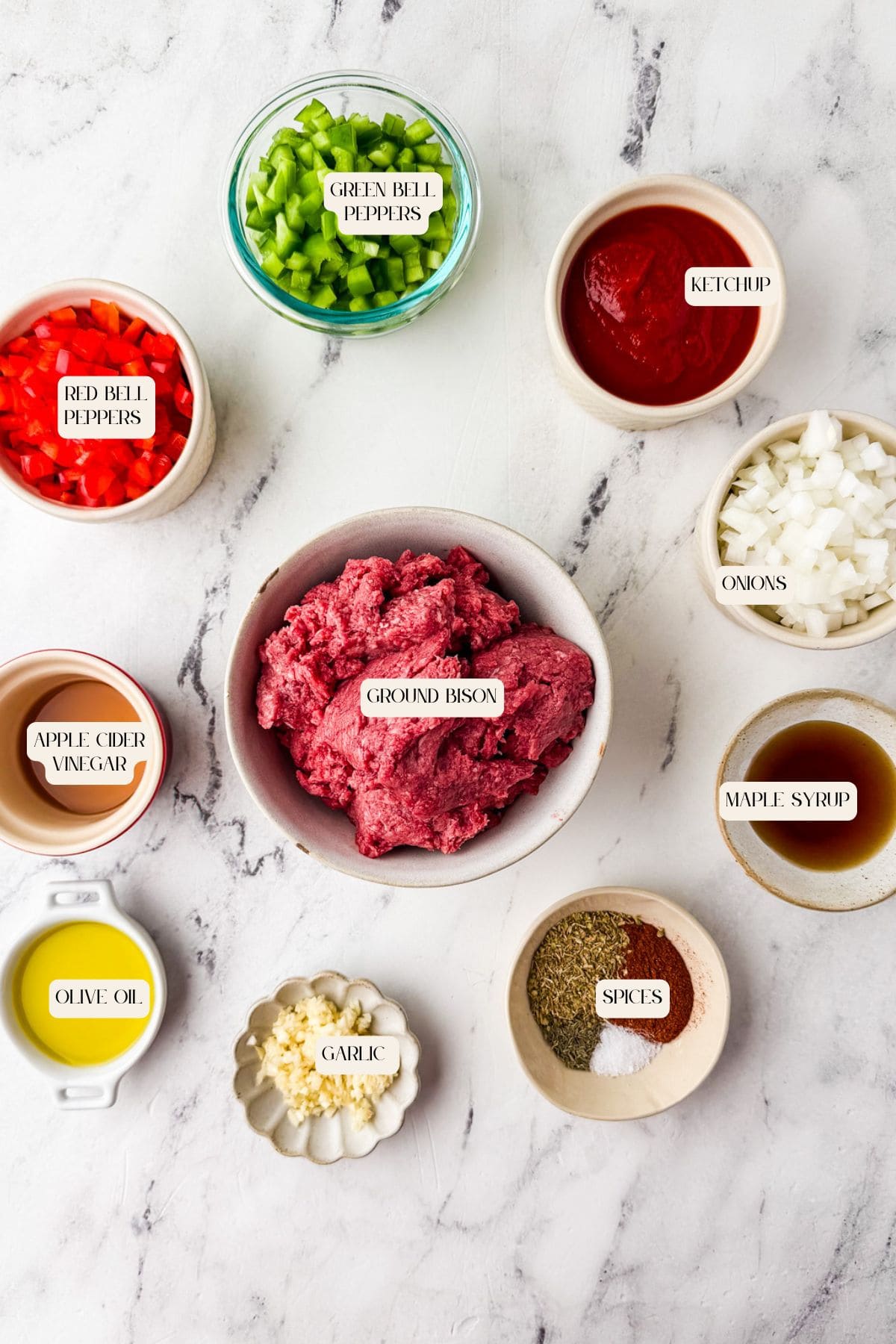 Ingredients for bison sloppy joe's are arranged on a white marble surface. Bowls hold ground bison, chopped green and red bell peppers, ketchup, diced onions, apple cider vinegar, maple syrup, olive oil, minced garlic, and spices.