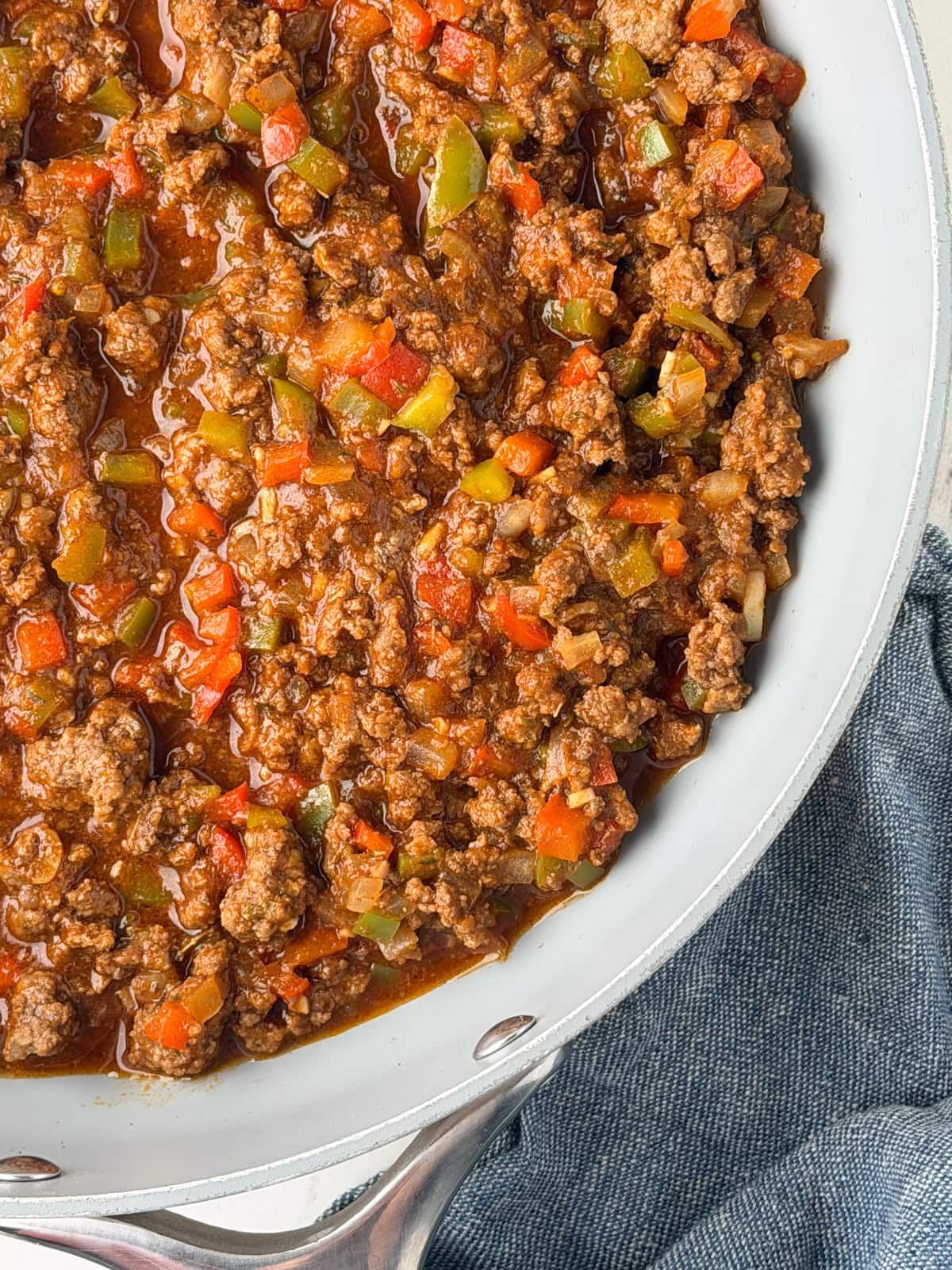 A skillet filled with cooked ground beef mixed with diced red and green bell peppers, onions, and a tomato-based sauce, resting on a blue cloth.