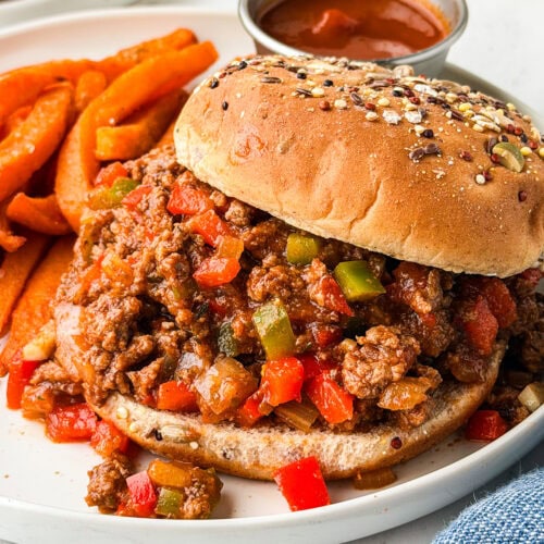 A sloppy joe sandwich with a whole grain bun, filled with ground beef, red and green bell peppers, and sauce, served on a white plate with sweet potato fries and a small cup of ketchup.
