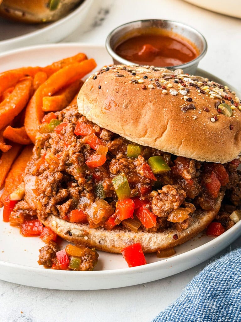 A sloppy joe sandwich with a whole grain bun, filled with ground beef, red and green bell peppers, and sauce, served on a white plate with sweet potato fries and a small cup of ketchup.