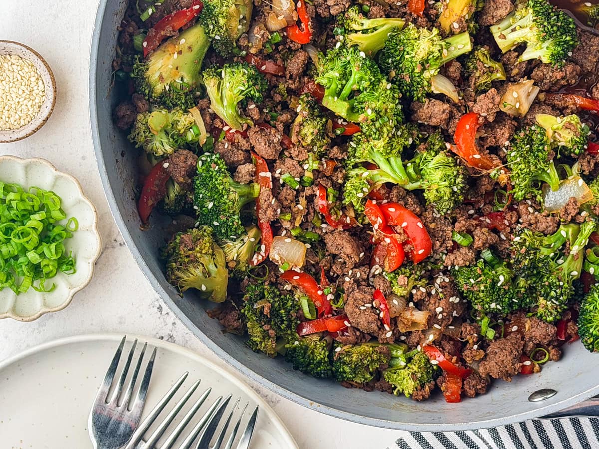 A skillet filled with a colorful stir-fry of ground beef, broccoli, red bell peppers, and onions, topped with sesame seeds. Next to it are small dishes of sliced green onions and sesame seeds, plus several forks on a plate.
