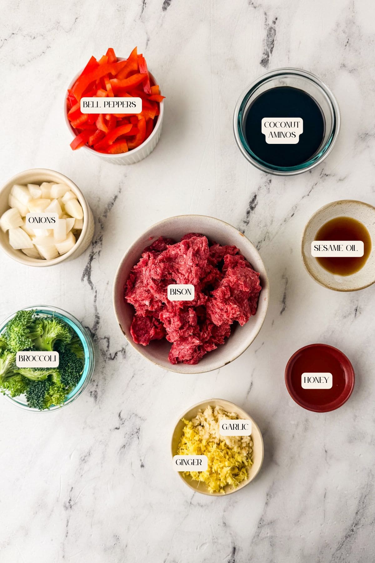 Bowls of raw ingredients arranged on a marble surface, including chopped bell peppers, onions, broccoli, and ground bison meat—perfect for a bison stir fry—plus coconut aminos, sesame oil, honey, minced ginger, and garlic. Each is labeled with text.