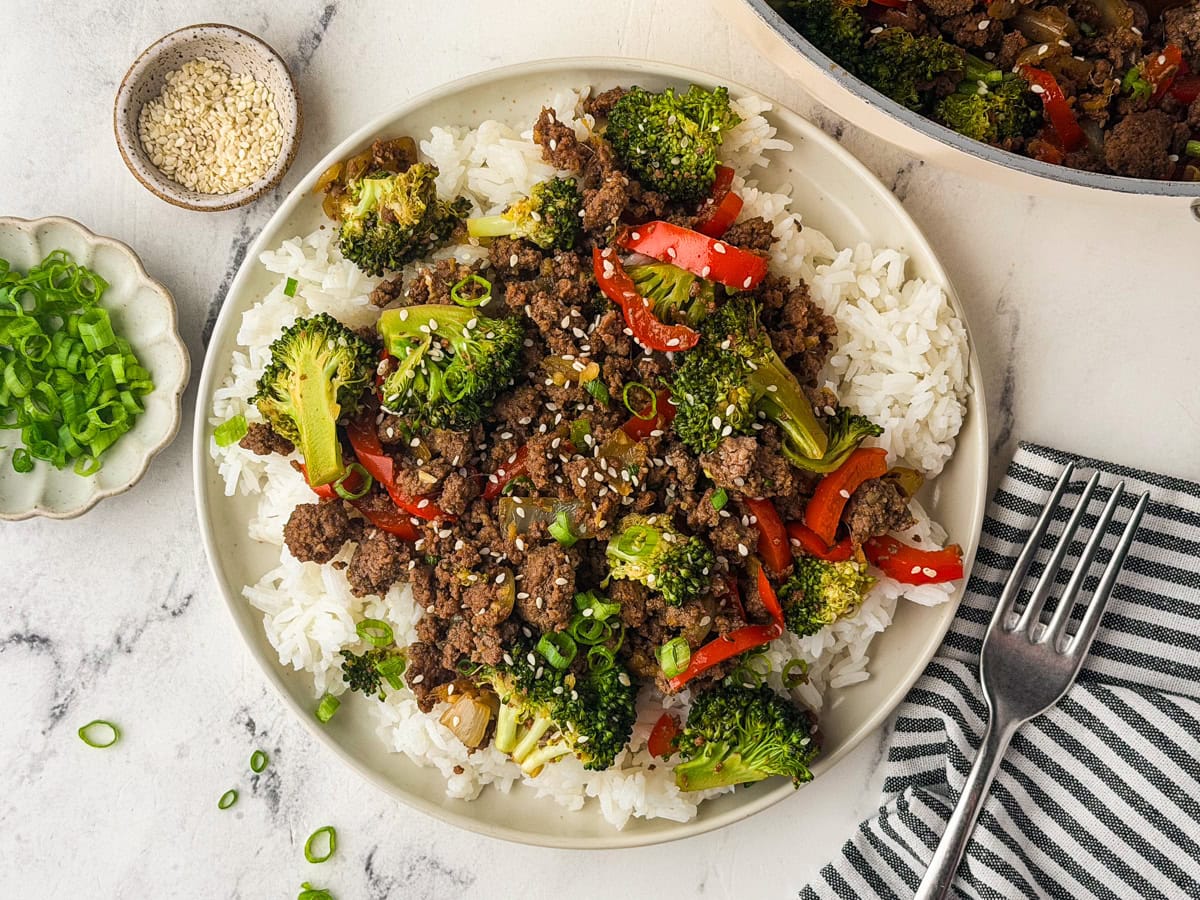 A plate of white rice topped with ground beef, broccoli, and red bell peppers, garnished with sesame seeds and green onions. A fork, striped napkin, sesame seeds, and chopped green onions are beside the plate.