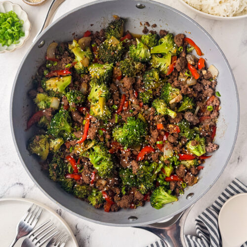 A skillet filled with a bison stir fry of broccoli, red bell peppers, and onions, sprinkled with sesame seeds. Plates, forks, white rice, green onions, and a small bowl of sesame seeds are nearby on a white surface.
