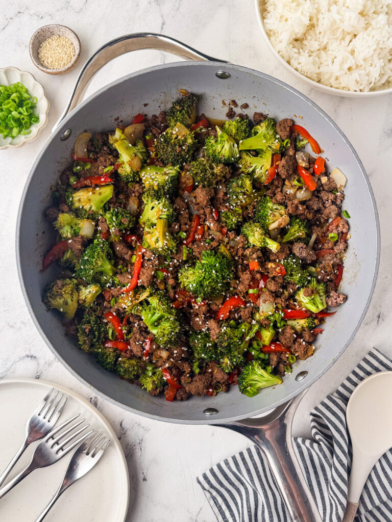 A skillet filled with a bison stir fry of broccoli, red bell peppers, and onions, sprinkled with sesame seeds. Plates, forks, white rice, green onions, and a small bowl of sesame seeds are nearby on a white surface.