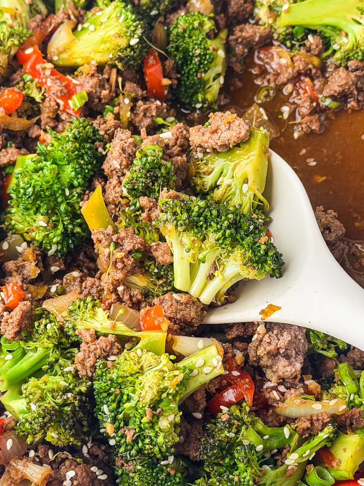 A close-up of a stir-fry with ground beef, broccoli florets, red bell peppers, and sesame seeds, served with a white spoon in a savory sauce.