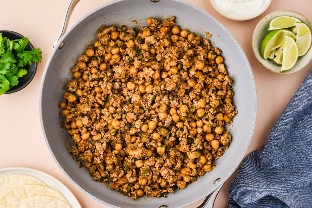 A skillet filled with cooked ground meat and chickpeas, perfect for ground chicken tacos, is surrounded by a bowl of cilantro, lime wedges, tortillas, and a blue napkin on a light-colored surface.