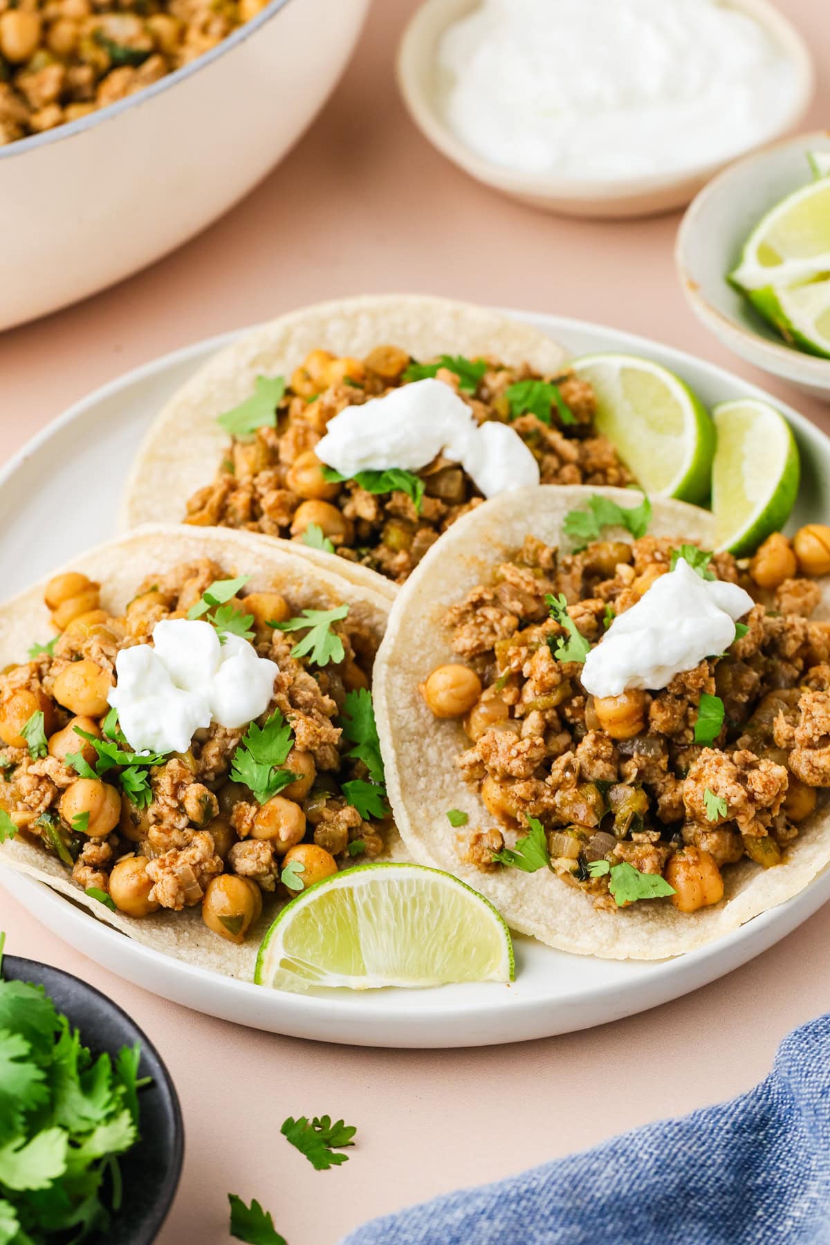 Three corn tortillas filled with a chickpea and ground chicken taco mixture, topped with dollops of sour cream and fresh cilantro, served on a plate with lime wedges. Bowls of toppings and herbs are in the background.