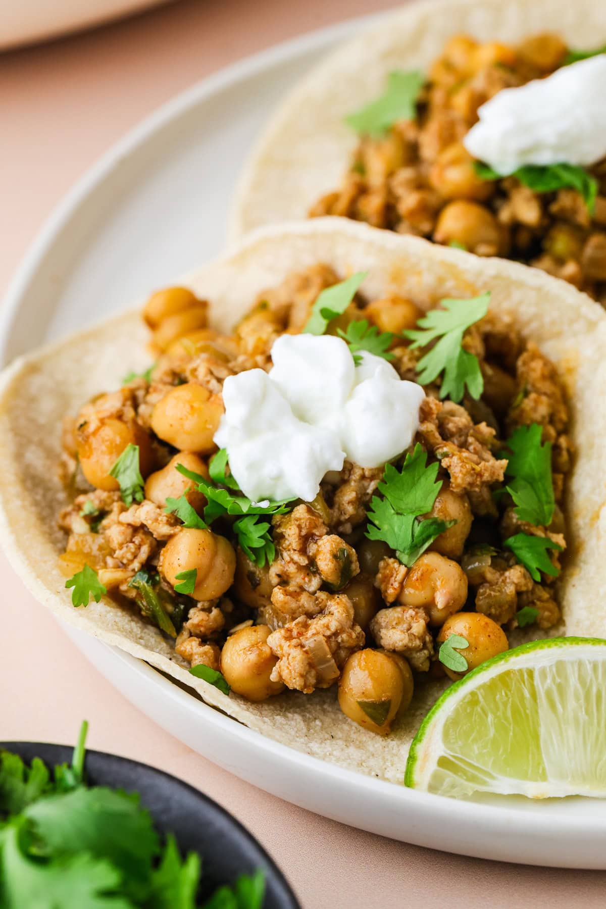 Two soft tortillas filled with spiced chickpeas, ground chicken tacos, fresh cilantro, and a dollop of sour cream, served on a white plate with a lime wedge on the side.