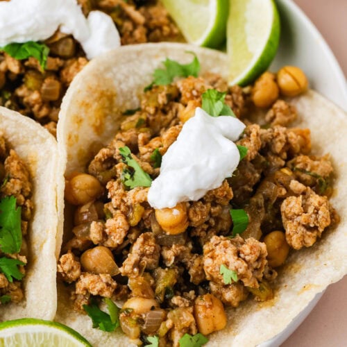 Three soft tacos filled with seasoned ground chicken tacos, chickpeas, and chopped herbs, topped with sour cream, are served on a plate with fresh lime wedges.