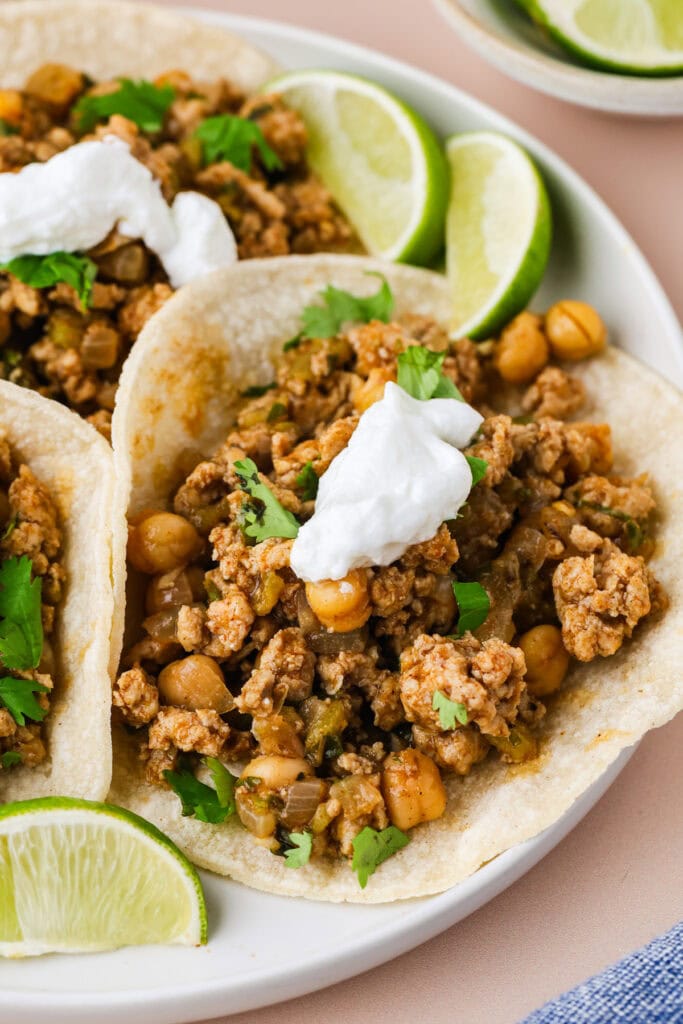 Three soft tacos filled with seasoned ground chicken tacos, chickpeas, and chopped herbs, topped with sour cream, are served on a plate with fresh lime wedges.