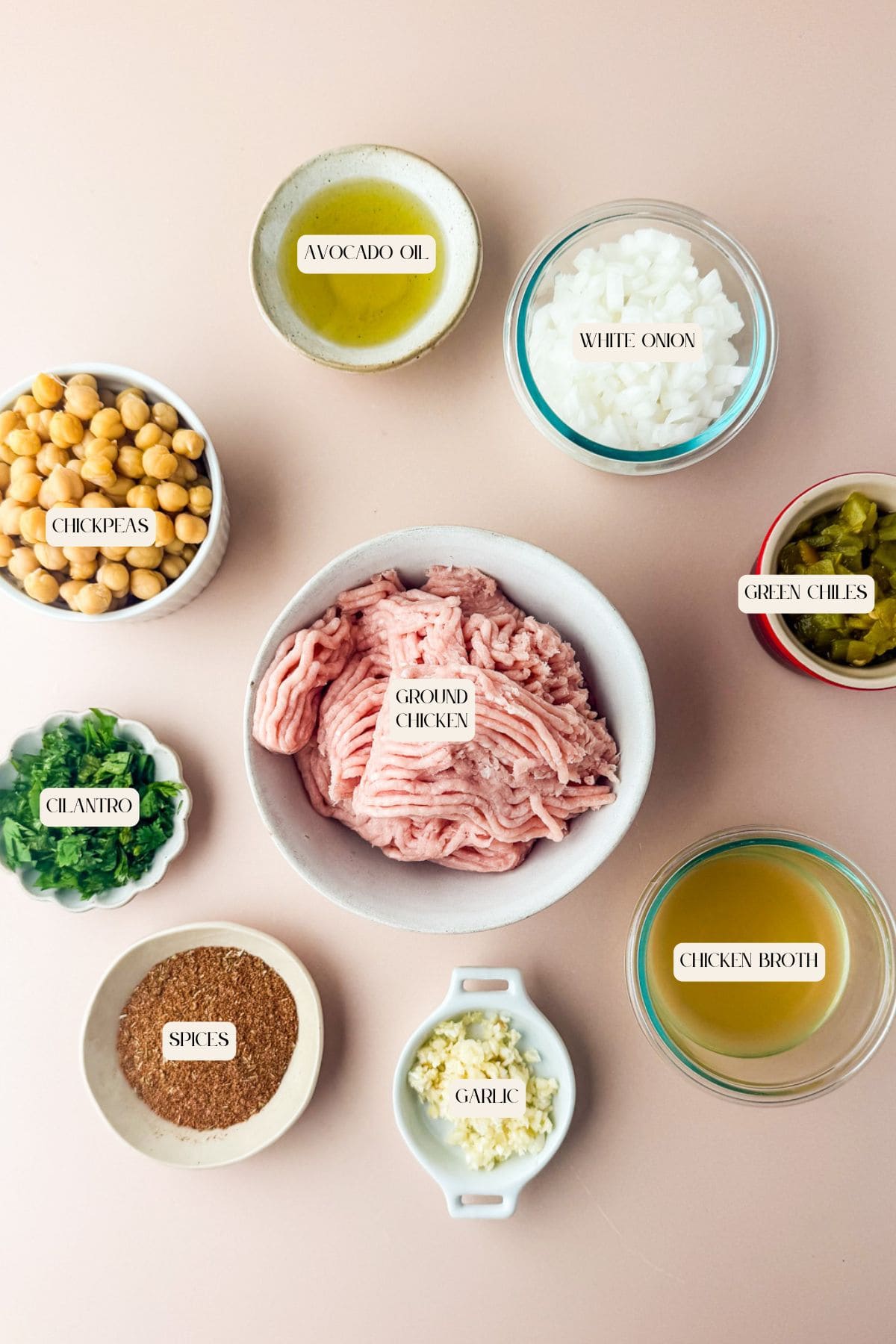 Overhead view of labeled bowls containing ingredients for ground chicken tacos, including ground chicken, chickpeas, avocado oil, white onion, green chiles, chicken broth, garlic, spices, and chopped cilantro on a light pink background.
