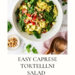 A bowl of Caprese Tortellini Pasta Salad with spinach, cherry tomatoes, mozzarella, walnuts, and basil sits on a white surface, surrounded by fresh salad ingredients, a wooden spoon, and utensils. Text reads Easy Caprese Tortellini Salad.