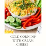A bowl of cold corn dip with cream cheese, topped with shredded cheese and herbs, is surrounded by sliced cucumbers, red bell pepper, celery sticks, and tortilla chips on a platter.