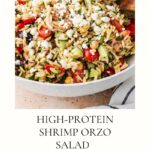 A bowl of Shrimp Orzo Salad with vegetables, herbs, and crumbled cheese, mixed and served with wooden utensils. Recipe title High-Protein Shrimp Orzo Salad is shown below the image.