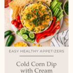A bowl of delicious corn dip with cream cheese, topped with fresh herbs and shredded cheese, is surrounded by crispy chips, celery sticks, cherry tomatoes, jalapeños, lime slices, and sweet corn.