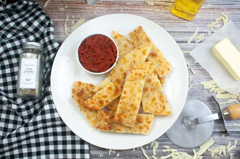 A white plate with cheesy breadsticks and a small bowl of marinara sauce, surrounded by shredded cheese, a spice jar, a stick of butter, a pizza cutter, and a checkered napkin on a wooden table.